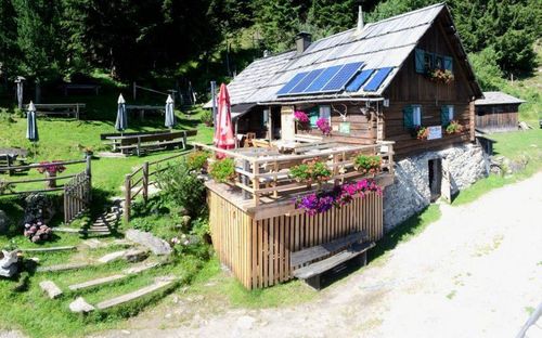 Ein rustikales Blockhaus mit Solarmodulen auf dem Dach ist von üppigem Grün umgeben, mit einer Holzterrasse und einer Bank in der Nähe.