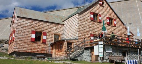 Ein Holzhaus mit rostigen Dach, Holzläden und einer Holzterrasse mit einer sitzenden Person. Es gibt zwei Bänke, einen Besen und einen geschlossenen Regenschirm.