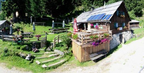 Ein Luftbild eines kleinen Holzhauses mit Solarpanelen auf dem Dach, umgeben von einem grünen Rasen, mit einem Holzzaun und einer Holzterrasse mit Blumen und Pflanzen.