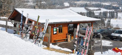 Eine Holzhütte mit einem schneebedeckten Metalldach. Vor ihr sind mehrere Skier und Stöcke aufgestellt. Im Hintergrund befindet sich eine schneebedeckte Landschaft mit Bäumen.