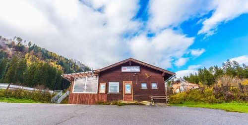 Ein kleines Holzgebäude namens 'Minnedorfer Hütte' steht vor dem Hintergrund eines Berges mit üppigen Bäumen und einer Bank davor.