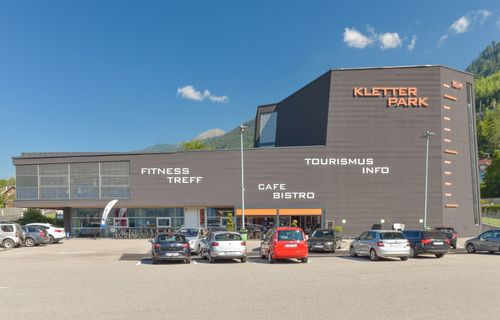 Die Vorderseite des Kletterpark-Gebäudes hat Schilder für Fitness Treff, Tourismus Info und Cafe Bistro. Autos sind draußen geparkt.