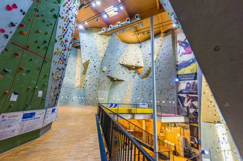 Ein Indoor-Kletterzentrum mit mehreren Kletterwänden. Helle Beleuchtung und verschiedenfarbige Griffe sind zu sehen. Eine Treppe führt zu einer Galerie.