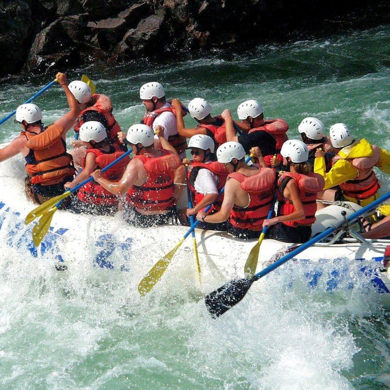 Eine Gruppe von Menschen treibt auf einem Fluss, paddelt und trägt Sicherheitswesten. Das Wasser spritzt um sie herum.