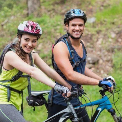 Zwei Personen auf Mountainbikes lächeln für ein Foto. Eine Frau im grünen Tanktop und ein Mann im blauen Tanktop tragen beide Helme. Sie sind von grünem Gras und Felsen umgeben.
