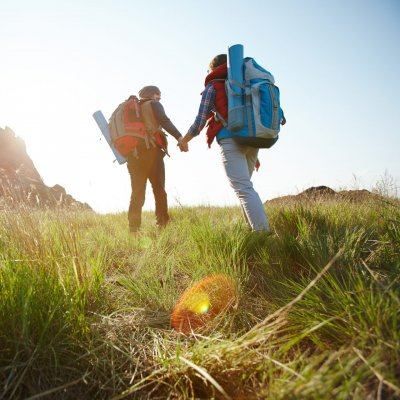 Zwei Wanderer, einer mit rotem und der andere mit blauem Rucksack, gehen Hand in Hand auf einem Grasfeld unter einem klaren Himmel.