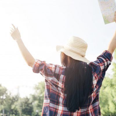 Eine Frau mit langen Haaren steht mit erhobenen Armen, hält eine Karte in der Hand, trägt einen Hut und ein kariertes Hemd in einem sonnigen Außenbereich.