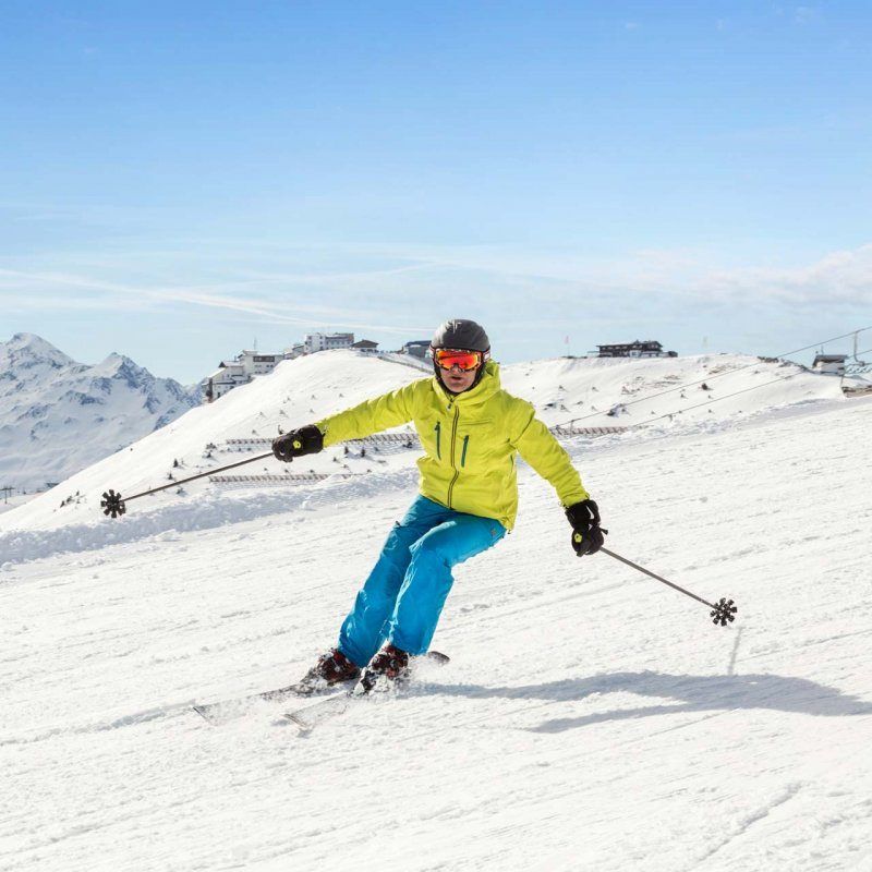 Ein Skifahrer in einem leuchtend gelben Jackett und blauen Hosen gleitet den schneebedeckten Hang hinunter, unter einem klaren Himmel. Im Hintergrund sind Berge und Gebäude sichtbar.