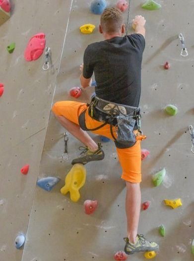 Ein Mann klettert an einer Kletterwand, trägt orange Shorts und Turnschuhe und hat verschiedene farbige Griffe angebracht.