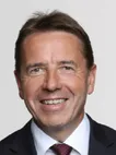 Ein lächelnder Mann in formeller Kleidung, möglicherweise ein Geschäftsmann, steht vor einem weißen Hintergrund.