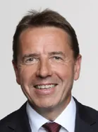 Ein Mann mit kurzen Haaren, der einen schwarzen Anzug und eine rote Krawatte trägt. Er lächelt und posiert wahrscheinlich für ein Foto.