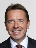 Ein Mann mit kurzen Haaren, der einen schwarzen Anzug und eine rote Krawatte trägt. Er lächelt und posiert wahrscheinlich für ein Foto.