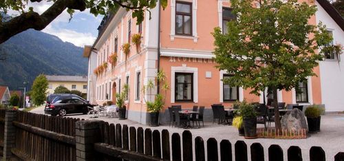 Ein rosafarbenes Gebäude mit mehreren Fenstern hat Tische und Stühle im Freien unter einem Baum. Das Gebäude hat ein Schild, das 'Restaurant' steht. Ein Auto ist vor dem Gebäude geparkt.