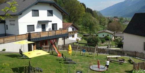 Ein Luftbild eines Hauses mit einem großen Garten. Der Garten hat Schaukel, Rutsche und Spielgeräte. Zwei Kinder spielen im Garten. Es gibt auch Sonnenschirme und Liegestühle. In der Umgebung befinden sich weitere Häuser, und Berge sind in der Ferne.