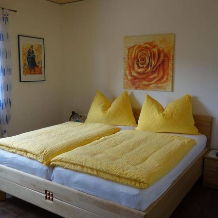 Ein Schlafzimmer mit einem Doppelbett, gelben Kissen und zwei Gemälden an der Wand. Ein Gemälde zeigt eine Rose, das andere ein Porträt.