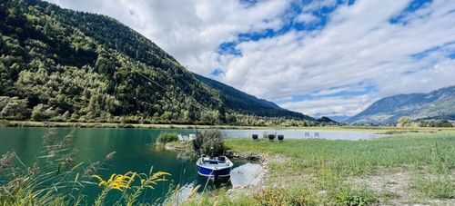 Ein kleines blau-weißes Boot am Ufer eines Sees mit Bergen und ein paar Stühlen im Hintergrund.