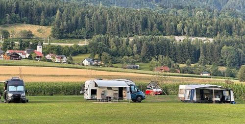 Mehrere Wohnmobile auf einem grünen Feld geparkt. Im Hintergrund ein kleines Dorf mit Häusern und Bäumen. Der Himmel ist klar und sonnig.