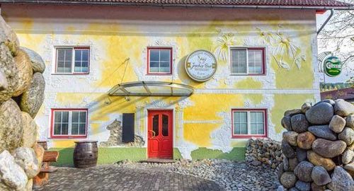 Ein gelbes und grünes Gebäude mit einer roten Tür und einem Fass daneben, umgeben von Steinhaufen. Das Gebäude hat ein rundes Schild mit dem Text 'Joschi Bar' über der Tür.