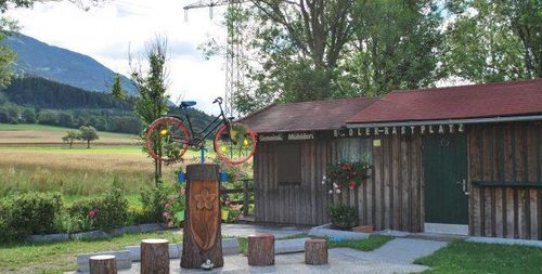 Ein Holzgebäude mit einem Fahrrad auf einem Pfosten und Blumen davor. Im Hintergrund sind Berge und ein Feld zu sehen.