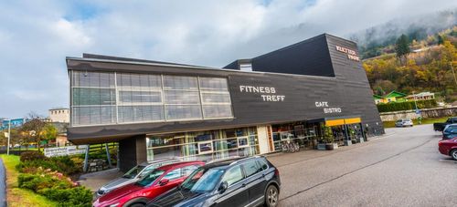 Die Vorderseite des Fitness Treff Gebäudes mit einem Café Bistro, mit großen Fenstern und geparkten Autos auf der Straße.