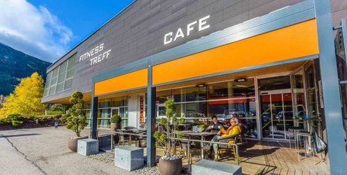 Ein Outdoor-Café namens Fitness Treff, wo zwei Personen draußen an Tischen sitzen, mit Bergen im Hintergrund.