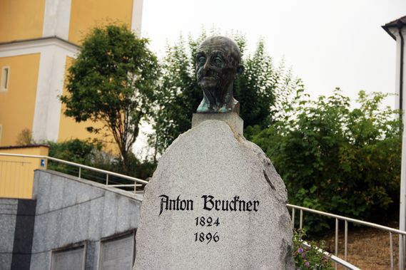 Eine Bronzebüste von Anton Brudhner, geboren 1824 und gestorben 1896, ist auf einer Steinplatte montiert, mit einem gelben Gebäude und Grünpflanzen im Hintergrund.