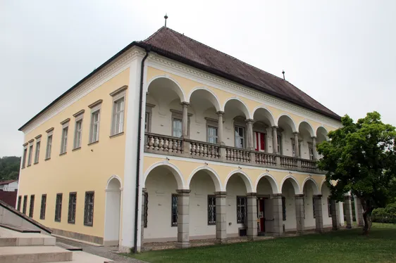 Ein großes gelbes Gebäude mit Rundbogenfenstern und Säulen, einem braunen Dach und einem Balkon im zweiten Stock.
