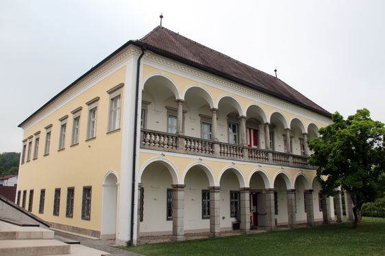 Ein großes gelbes Gebäude mit Rundbogenfenstern und Säulen, einem braunen Dach und einem Balkon im zweiten Stock.