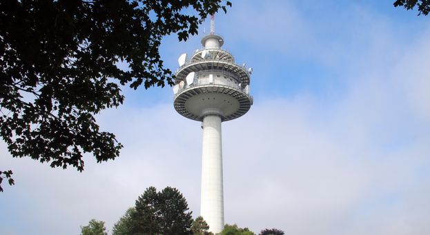 Ein hoher weißer Turm mit mehreren Satellitenschüsseln auf der Spitze, umgeben von Bäumen und blauem Himmel.