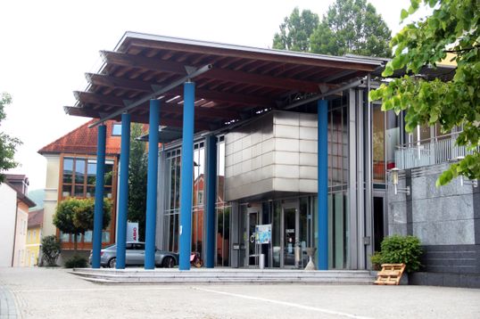 Ein modernes Gebäude mit einem Holzdach und blauen Säulen, mit Glaswänden und einem vor dem Gebäude geparkten Auto.