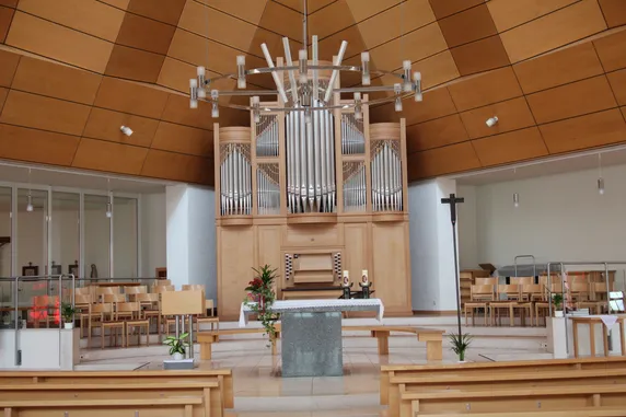 Ein leerer Kircheninnenraum mit einer großen Orgel, einem Kreuz und Kerzen auf dem Altar. Holzbänke und Pflanzen schmücken den Raum.