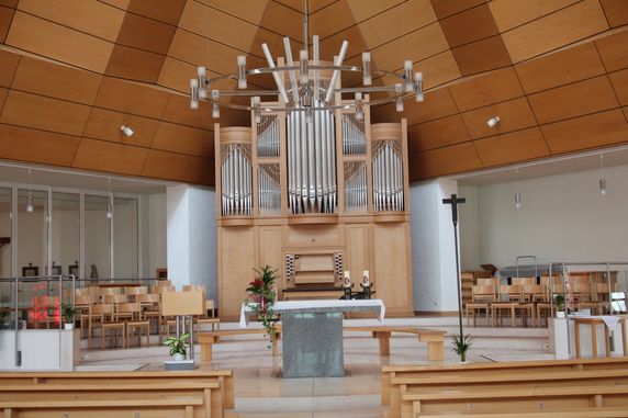 Ein leerer Kircheninnenraum mit einer großen Orgel, einem Kreuz und Kerzen auf dem Altar. Holzbänke und Pflanzen schmücken den Raum.