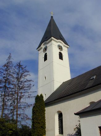 Ein hoher weißer Kirchturm mit schwarzem Dach und zwei Uhren, umgeben von Bäumen.