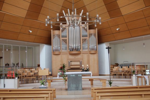 Das Innere einer modernen Kirche mit einer großen Orgel im Hintergrund, einem Tisch mit Blumen und Kerzen davor und hölzernen Kirchenbänken auf beiden Seiten.
