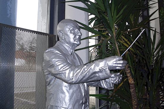 Eine silberne Statue eines Mannes in einem Anzug, der einen Taktstock hält, neben einer großen Pflanze.