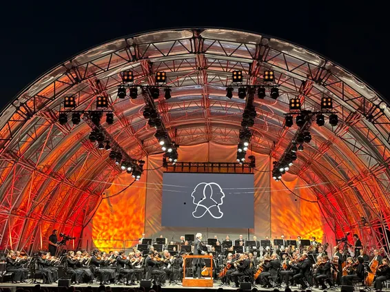 Ein Symphonieorchester spielt auf einer Bühne unter einem großen Bogen mit einer großen Leinwand, die ein Logo zeigt. Die Bühne ist mit roten und orangefarbenen Lichtern beleuchtet.