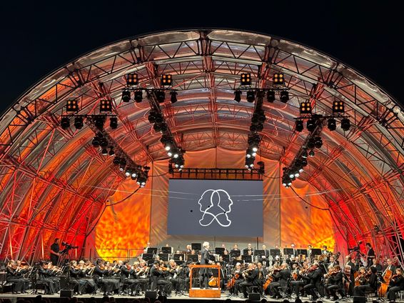 Ein Symphonieorchester spielt auf einer Bühne unter einem großen Bogen mit einer großen Leinwand, die ein Logo zeigt. Die Bühne ist mit roten und orangefarbenen Lichtern beleuchtet.