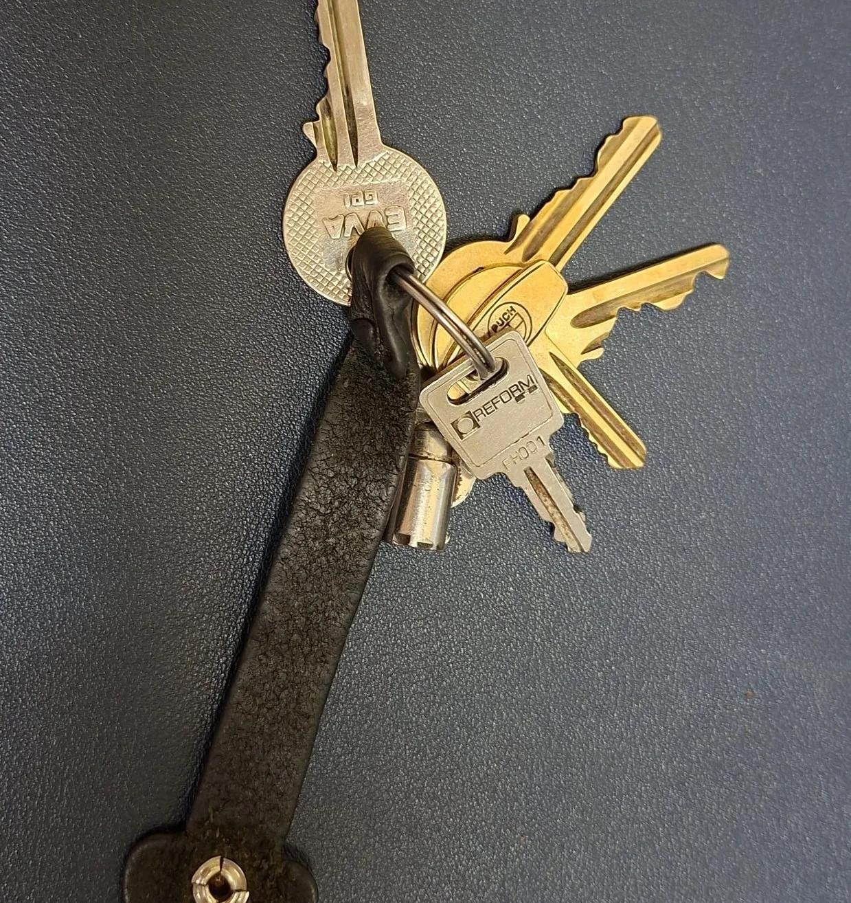 Eine Sammlung von goldenen Schlüsseln mit einem schwarzen Lederschlüsselring auf einer grauen Oberfläche.