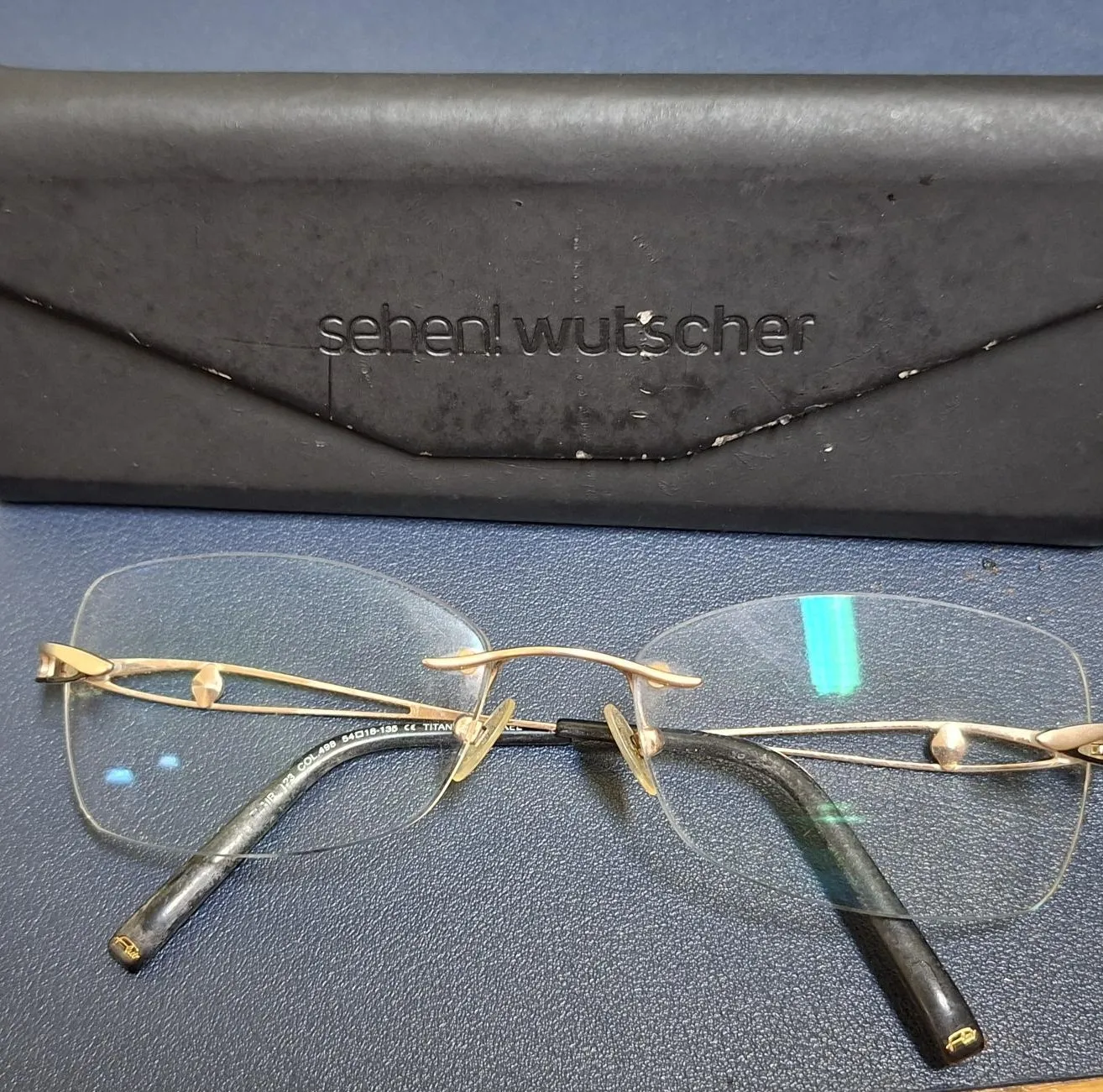 Ein Paar Brille mit goldfarbenen Rahmen und klaren Gläsern liegt auf einer blauen Oberfläche. Hinter der Brille befindet sich eine schwarze Ledertasche mit dem Text 'sehen! wutscher' darauf.
