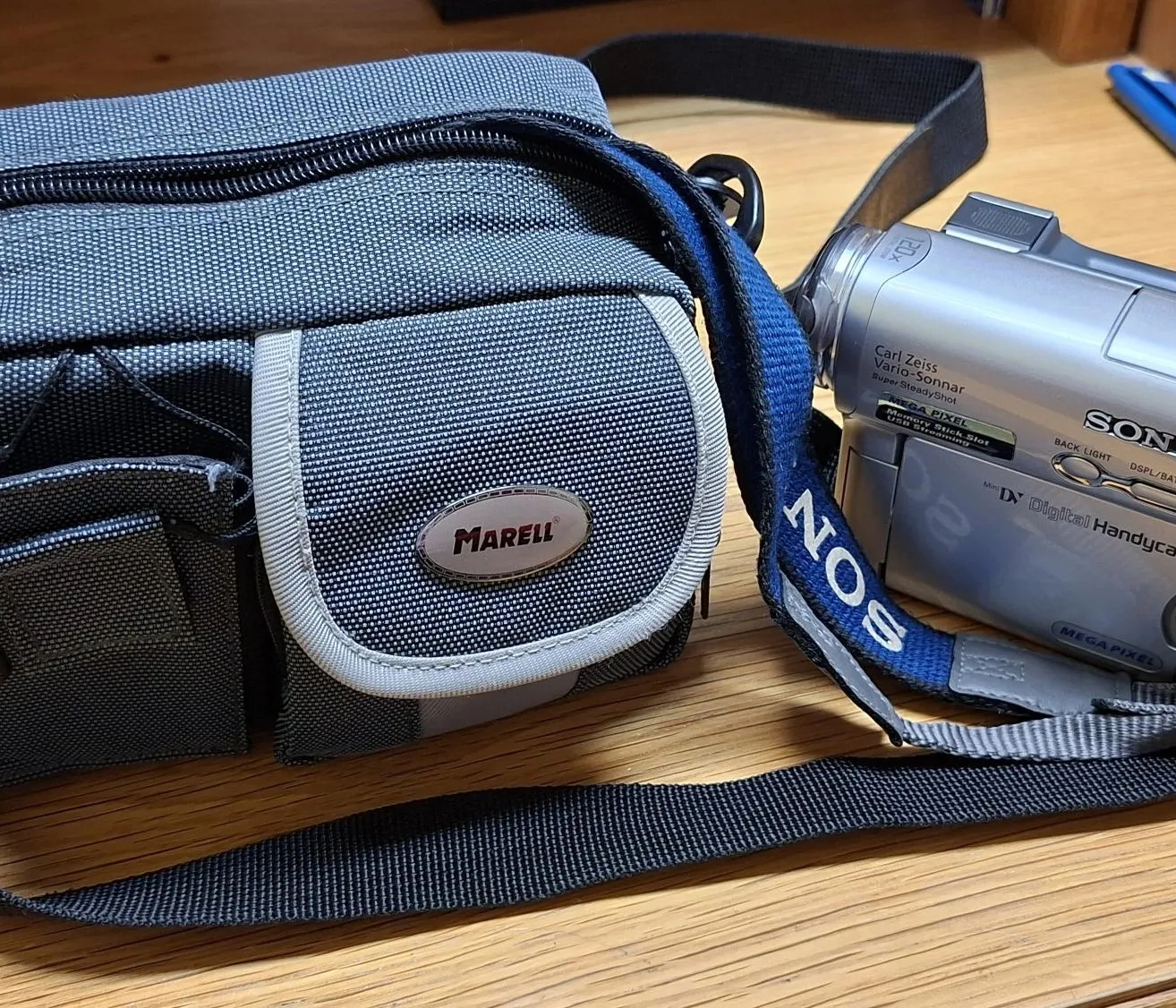 Eine Sony-Kamera mit einem Gurt liegt auf einem Holztisch. Die Kamera hat ein Markenlogo an der Seite und eine Tasche vorne.