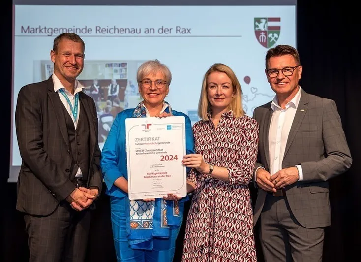 Drei Personen stehen lächelnd auf einer Bühne. Eine Frau hält ein Zertifikat. Hinter ihnen zeigt eine Leinwand 'Marktgemeinde Reichenau an der Rax' und ein Logo.