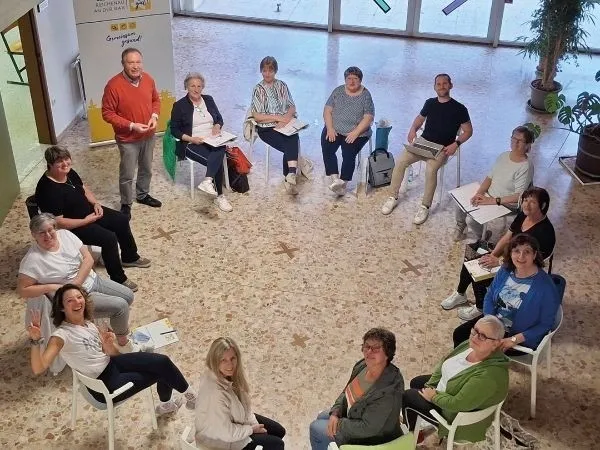 Eine Gruppe von Erwachsenen, sowohl Männer als auch Frauen, sitzen in einem Raum in einem Kreis, wahrscheinlich eine Besprechung. Sie haben Papiere und Taschen bei sich. Ein Mann steht vor ihnen und schaut sie an. Es gibt Topfpflanzen im Raum.