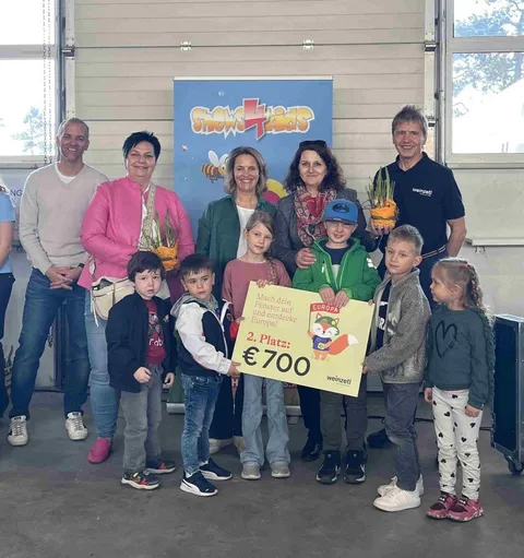 Eine Gruppe von Erwachsenen und Kindern steht zusammen, einige halten Blumen und ein gelbes Poster mit den Worten '2. Platz' und '700'. Hinter ihnen befindet sich ein Schild mit den Worten 'Shows4Kids'.