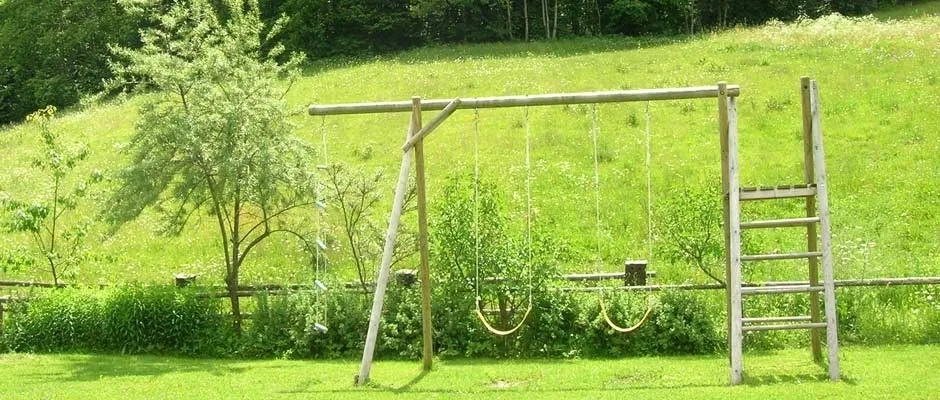 Ein Outdoor-Spielbereich mit zwei Schaukeln, die an einem Holzrahmen hängen, und einer Holzleiter auf der rechten Seite.