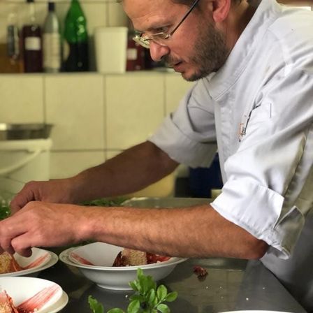 Ein Mann in einer weißen Kochuniform bereitet ein Gericht mit roter Sauce auf einem Arbeitstisch zu.