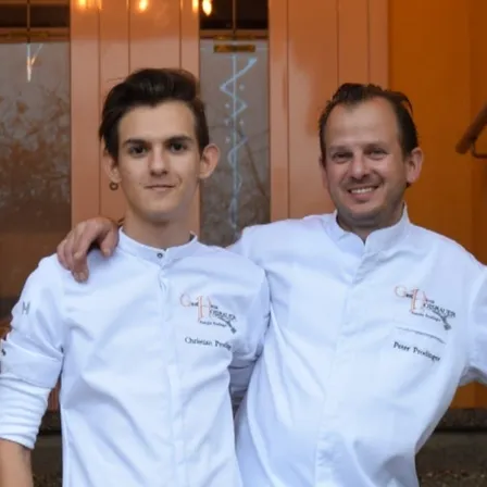 Zwei Köche, Christian und Peter, lächeln und posieren für ein Foto. Beide tragen weiße Kochuniformen mit gestickten Logos. Sie stehen eng beieinander.