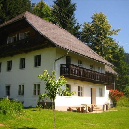 Ein traditionelles Haus mit einem braunen Dach und weißen Wänden, umgeben von einem üppigen grünen Rasen, mit einer Terrasse und einem Sitzbereich im Freien, vor dem Hintergrund von Bäumen und Bergen.
