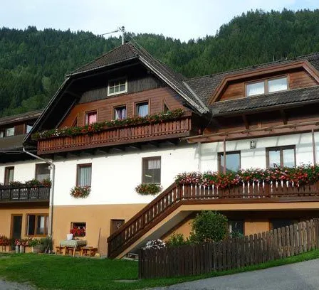 Ein zweistöckiges Haus mit einem braunen Dach, weißen Wänden und einem Holzzaun vorne. Das Haus hat einen Balkon mit roten Blumen im zweiten Stock.