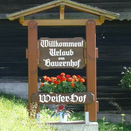 Ein hölzerner Schild vor einem Gebäude mit der Aufschrift 'Willkommen! Uelaub am Bauernhof.' Ein Blumenarrangement befindet sich auf dem Schild.