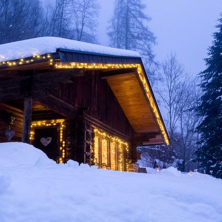 Ein kleines Holzhaus im Schnee, mit Lichtern auf dem Dach und den Fenstern, umgeben von Bäumen und einer herzförmigen Dekoration an der Tür.